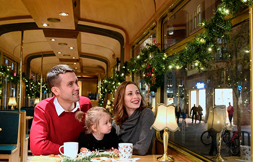 Mann, Frau und Tochter sitzen im weihnachtlich geschmückten historischen Fahrzeug. Vor ihnen stehen Tassen und Gebäck.