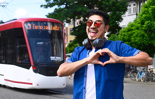 Junger Mann mit Sonnenbrille steht vor einer Straßenbahn und formt mit den Händen ein Herz.