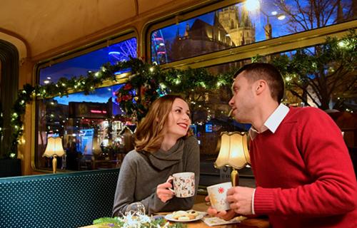 Mann und Frau sitzen im weihnachtlich geschmückten historischen Fahrzeug. Vor ihnen stehen Tassen und Gebäck. Im Hintergrund der weihnachtliche Domplatz.