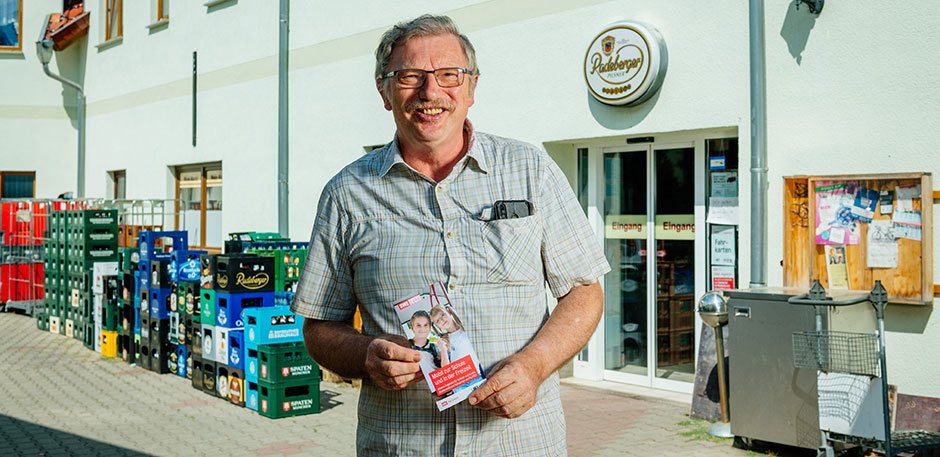 Mann steht mit Flyern in der Hand vor dem Eingang eines Dorfladens.