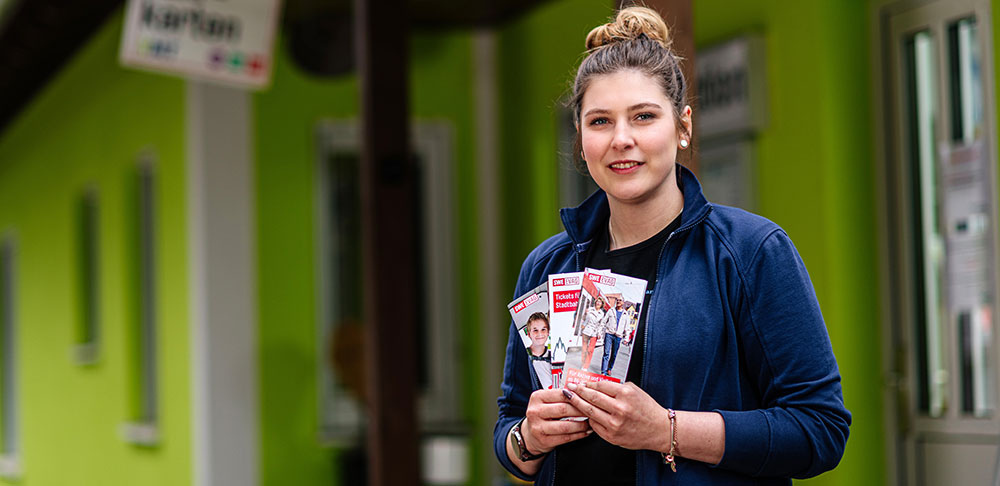 Eine Frau steht vor dem grün gestrichenen Empfangsgebäude eines Campingplatzes. In der Hand hält sie Fyer.