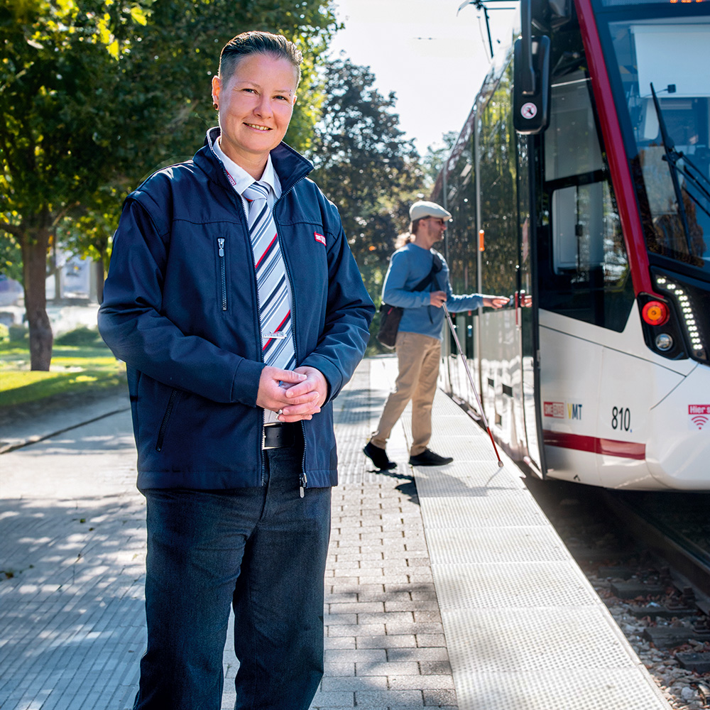 Fahrerin an der Haltestelle. Im Hintergrund eine Bahn und ein Mann mit Blindenstock auf dem Blindenleitstreifen.