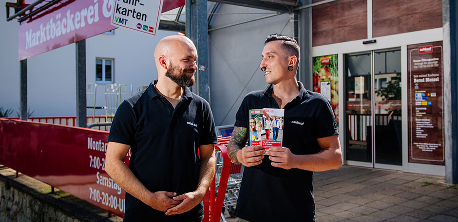 Zwei Männer stehen vor dem Eingang eines Supermarktes. Ein Mann hält Flyer in den Händen.