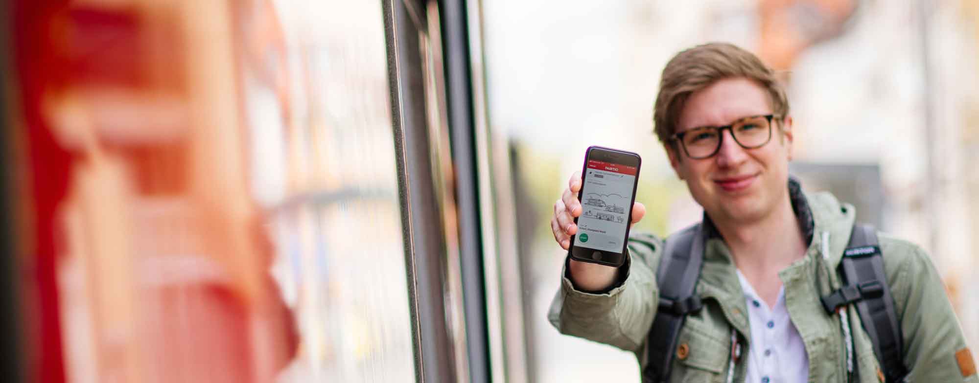 Fahrgast zeigt Handy mit FAIRTIQ-App auf dem Bildschirm. Rechts ist eine Straßenbahn zu sehen. 